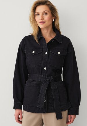 Vrouw draagt een zwart denimjack met knoopzakken op de borst en een gestrikte riem, staand tegen een effen lichte achtergrond.