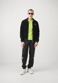 Man wearing black zip-up jacket, neon green shirt, black track pants with white and gray accents, white sneakers, and dark sunglasses standing on white floor.