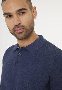 Námořnická modrá texturovaná polokošile s límečkem, která má fialové knoflíky a jemný vzor, zobrazená na horní části těla modelu.