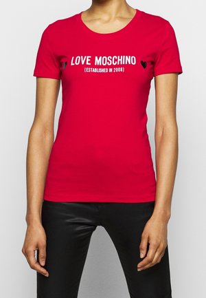 Frau trägt ein eng anliegendes rotes T-Shirt mit dem Text „LOVE MOSCHINO (ESTABLISHED IN 2008)“ und schwarzen Herzen, kombiniert mit schwarzen Hosen.