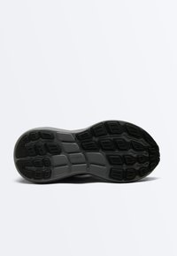 Semelle d'une chaussure de sport gris foncé avec un motif de bande de roulement texturé pour l'adhérence, présentée sur un fond clair uni.