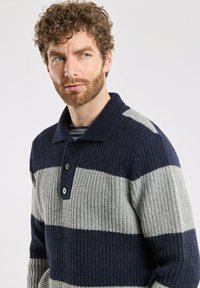 Pull en tricot avec des rayures bleu marine et grises, texture côtelée, col et patte de boutonnage à trois boutons. Présente un motif de rayures horizontales.