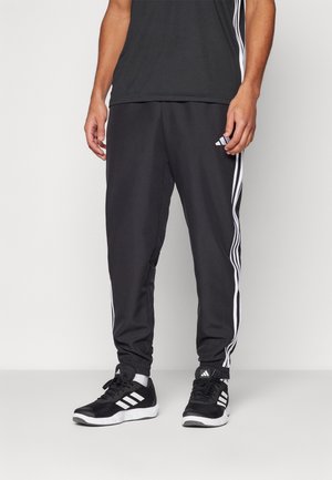 Mężczyzna w czarnych spodniach dresowych Adidas z białymi paskami oraz czarnych sneakersach Adidas, stoi na jednolitym tle.