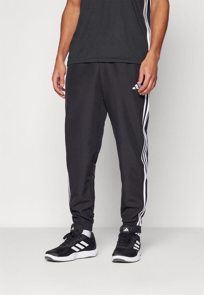 Homme portant un pantalon de survêtement Adidas noir avec des bandes blanches et des baskets Adidas noires, debout devant un fond uni.