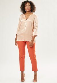 Satin-Pfirsich-Oberteil mit V-Ausschnitt und dreiviertellangen Ärmeln, kombiniert mit enganliegenden orangen Hose. Das Model trägt beigefarbene High Heels und goldene Ohrringe.