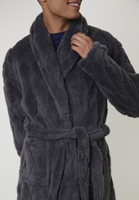 Persona sorridente che sistema il colletto di un accappatoio in peluche grigio scuro con colletto a scialle e cintura, mettendo in risalto la texture e il tessuto accogliente.