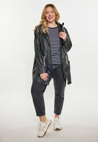 Black rubberized parka with hood, zip and snap closures, drawstrings, and side pockets, layered over a striped shirt and black jeans.