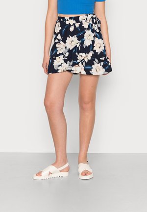 Skort floral com fundo azul-marinho, apresentando flores brancas e rosa claro, cintura ajustável e um design em camadas. Usado com sandálias brancas.