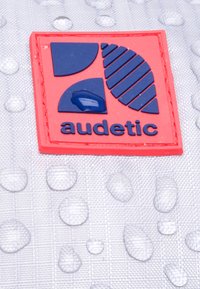 Röd fyrkantig logotyp med blå geometriska former och texten "audetic" på en strukturerad vit bakgrund med vattendroppar.