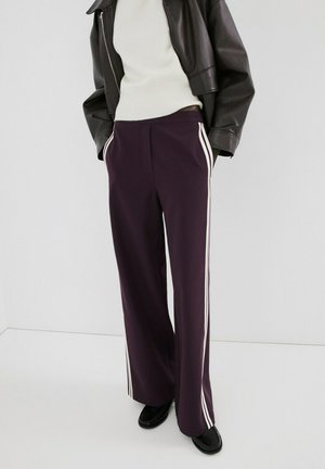 Persona vistiendo pantalones anchos de color púrpura oscuro con franjas blancas en los lados, suéter blanco, chaqueta de cuero negra y zapatos negros, de pie con las manos en los bolsillos.