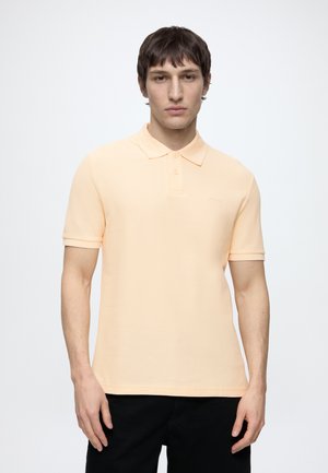 Ung mand med kort mørkt hår iført en lys ferskenfarvet kortærmet polo shirt og sorte bukser mod en ensartet hvid baggrund.