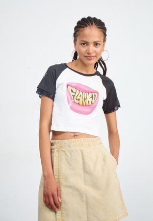 White cropped T-shirt with black sleeves featuring a graphic of pink lips and the word "FLACKY." Paired with a beige skirt.