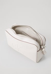 Bílá kosmetická taška se zipem s raženým logem, hladký kožený materiál, zaoblený tvar a kontrastní hnědé akcenty na okrajích.