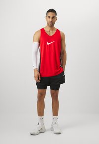 Camiseta sin mangas roja de Nike con un logo blanco, combinada con pantalones cortos negros. Una manga de brazo blanca y zapatillas blancas completan el look deportivo.