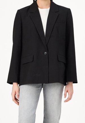 Personne portant un blazer noir à un seul bouton sur une chemise blanche et un jean gris clair, montrée du cou aux hanches sur un fond blanc.