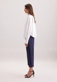 Blusa bianca, comoda e ampia con bottoni laterali, abbinata a pantaloni tapered blu navy e sandali neri con tacco. Tessuto liscio, design semplice e accenti minimali.