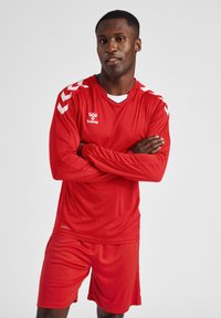 Rode sportoutfit bestaande uit een lange mouwen truitje en shorts. Het truitje heeft witte chevronaccenten op de mouwen en een merknaam op de voorkant.