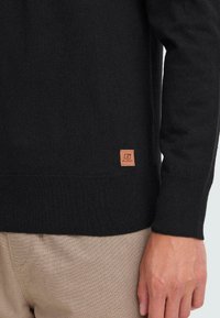 Close-up van een persoon die een zwarte trui draagt met een klein bruin "Blend" logo tag bij de zoom en beige gestructureerde broek.