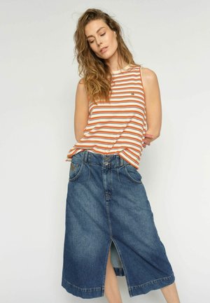 Kvinde med langt hår iført en orange og hvidstribet ærmeløs top og en blå denimnederdel til knæene med en frontslids.