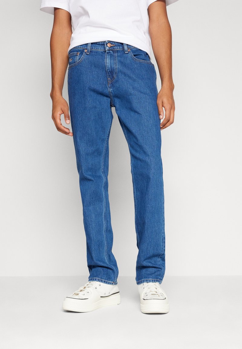 Vaqueros de mezclilla azul medio, con corte recto, diseño de cinco bolsillos y cierre con botón de latón, combinados con zapatillas de lona blancas.