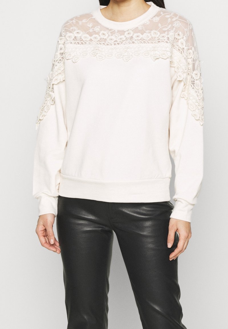 Licht crèmekleurig sweatshirt met kanten details op de schouders en borst. Getailleerde boorden, ronde hals, gemaakt van zacht materiaal, gecombineerd met zwarte leren broek.
