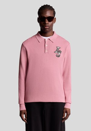 Långärmad rosa polo i strukturerad tyg med tre knappar och en broderad "LS"-logotyp på bröstet, i kombination med mörka byxor.