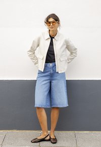 Femme portant des lunettes de soleil ambre, une veste zippée beige rayée, un haut noir, un short en jean longueur genou et des chaussures plates en mesh noir, debout contre un mur gris-blanc.
