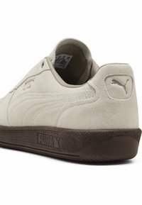 Adidași din suede bej cu o talpă din cauciuc maro, având un toc plat, un design minimalist și un logo Puma embosat pe toc.
