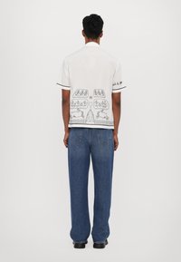 Camisa blanca de manga corta con diseños bordados en negro inspirados en la naturaleza en la parte posterior, combinada con unos pantalones vaqueros amplios de mezclilla azul.