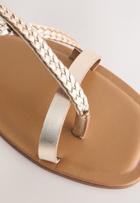 Sandali bassi con cinturino intrecciato in argento metallizzato e suola marrone liscia, dal design minimalista e punta aperta.