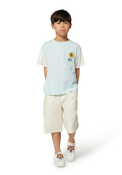 Maglietta azzurra chiara con maniche corte bianche e una grafica di girasole, abbinata a pantaloni corti di colore crema e sneakers bianche con un design di tigre.