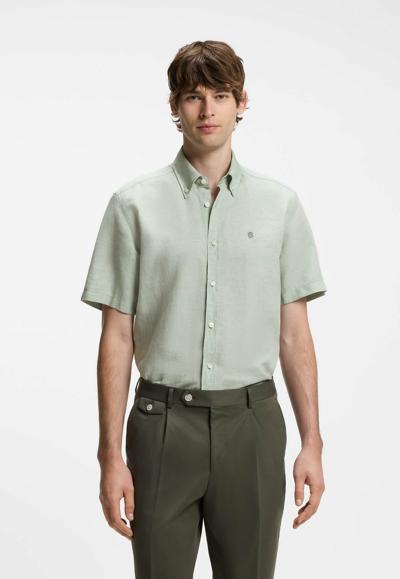 Jeune homme debout, portant une chemise verte claire à manches courtes boutonnée et un pantalon vert foncé sur un fond blanc uni.