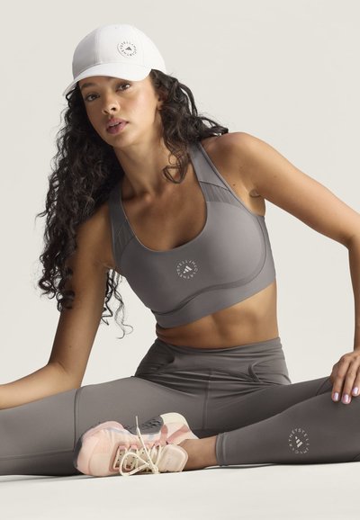 Woman in gray sports bra and leggings stretching on floor wearing white cap and light pink athletic shoes against plain background.