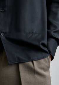 Camicia nera con bottoni realizzata in tessuto morbido, con un logo discreto vicino all'orlo. Dettaglio dei bottoni visibile sul lato sinistro.