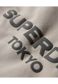 Tessuto grigio chiaro con grandi lettere in rilievo nere che formano la scritta "SUPERDRY TOKYO". La superficie ha una consistenza morbida e leggermente pelosa.