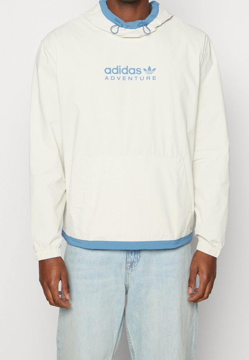Ljusgrön vindjacka med blå detaljer, utrustad med en huva med dragsko och en framficka i kängurustil. "adidas ADVENTURE"-logotypen visas.