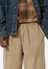 Džínová bunda s mosadznými cvokmi cez károvanú košeľu. Dolu béžové kordové nohavice s voľným strihom a bočnými vreckami s ribbovanou textúrou.