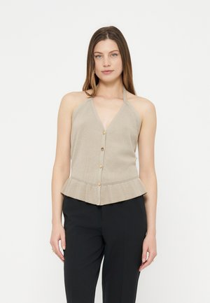 Femme portant un haut halter beige boutonné avec un ourlet volanté et un pantalon noir, debout sur un fond blanc uni.