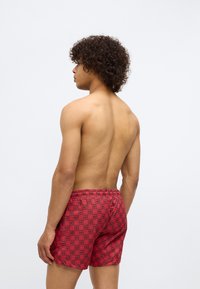 Red swim shorts featuring a grid pattern with alternating "HU" text. Elastic waistband and short-cut design. Smooth texture.