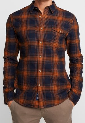 Mann trägt ein geknöpftes, langärmliges kariertes Hemd mit orange-blauen Karos, kombiniert mit beigen Hosen und hat die Hände in den Taschen.