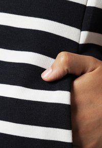 Niebiesko-białe paski dziane o gładkiej fakturze z delikatnym rozciągnięciem, ręka trzymająca krawędź. Paski są poziome.