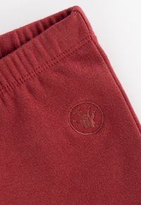 Red cotton sweatpants feature an elastic waistband and a small logo patch with a circular border, showcasing a soft, brushed texture.