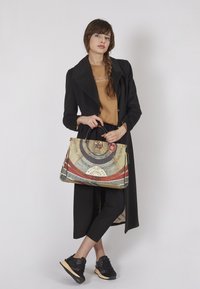 Donna in un lungo cappotto nero e pantaloni neri che tiene una grande borsa a tracolla con motivi circolari, in piedi davanti a uno sfondo chiaro uniforme.