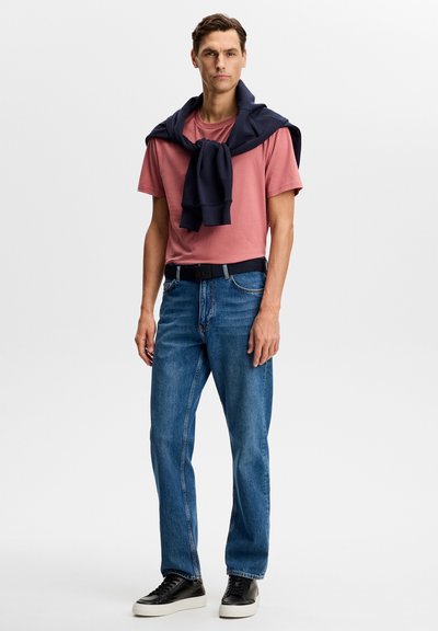 Homme portant un t-shirt rose, un jean bleu, une ceinture noire, des baskets noires, et un sweat-shirt bleu marine noué autour des épaules, se tenant contre un fond blanc.