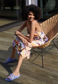 Colorful patterned dress in orange, pink, and purple, paired with light blue slide sandals. Braided wicker chair and wooden flooring visible.