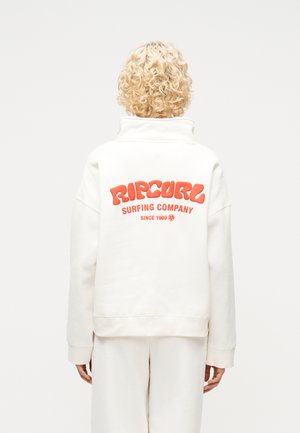 Osoba s kučeravými blond vlasmi v bielom pulóvri Rip Curl s oranžovým nápisom "Surfing Company Since 1969", zobrazená zozadu.