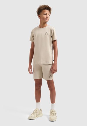 Jong persoon staand, gekleed in een bijpassend beige shirt met korte mouwen en korte broek, met witte sokken en beige sneakers, tegen een grijze achtergrond.