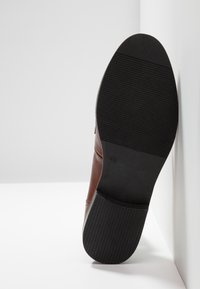 Chaussure en cuir marron avec semelle en caoutchouc noire orientée vers le haut, montrant le talon texturé et l'avant de la semelle sur fond blanc.