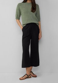 Femme portant un haut en tricot à manches courtes vert, un pantalon noir à jambes larges et cropped, et des sandales plates noires, debout devant un fond uni.