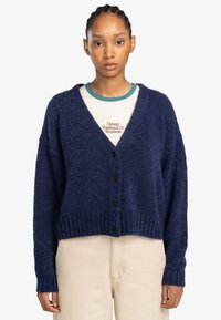 Marineblauwe fluffy cardigan met V-hals, lange mouwen en vijf zwarte knopen. Geribbelde zoom en boorden, gedragen over een crèmekleurig overhemd.